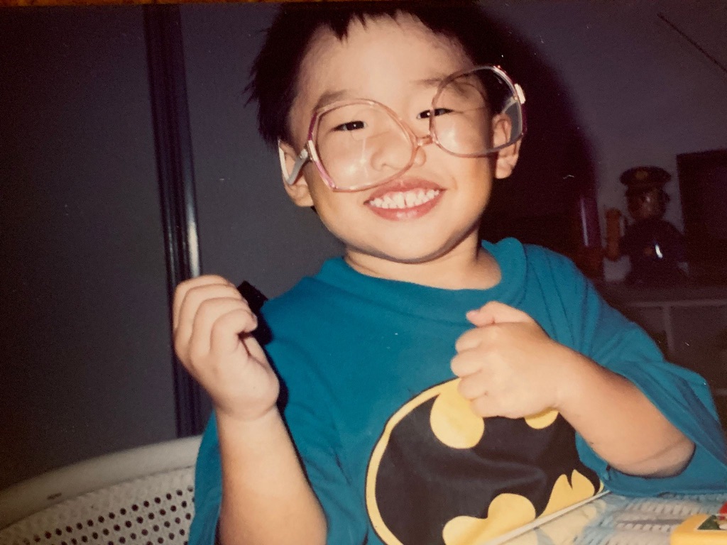 Bryan as a kid, grinning in oversized glasses and a Batman shirt.
